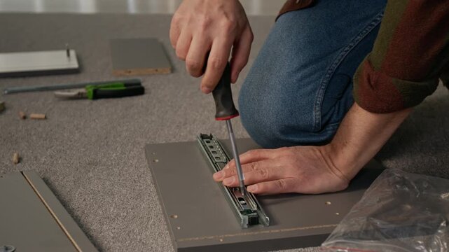Medium close-up of hands of anonymous man attaching runner to drawer panel while assembling flatpack furniture, tightening bolts with screwdriver on floor carpet, holding with knee
