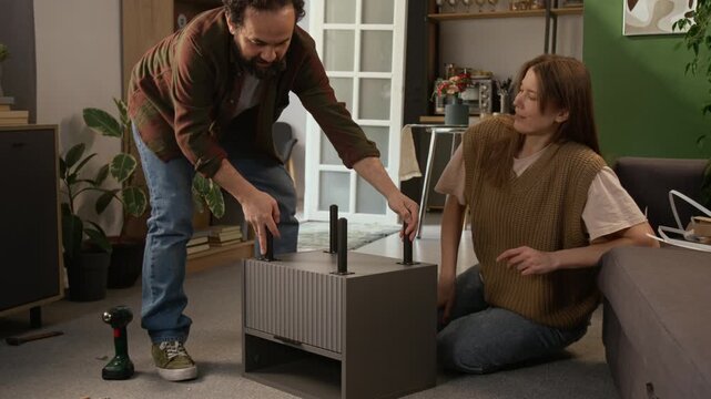 Full shot of joyous middle-aged Caucasian couple attaching legs to flat pack drawer, finishing with task, turning over, admiring work, then giving each other high fives, hugging and kissing