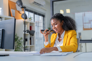 Confident professional businesswoman using smartphone voice message while reviewing documents at...