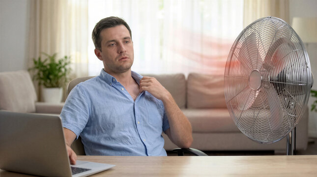 A warm man in a blue shirt tries to cool down with an electric fan in his living room while working on a laptop.
