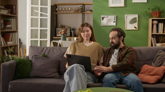 Medium full shot of mature Caucasian couple sitting together on couch in living room, viewing merchandise in online store on laptop, choosing and discussing new furniture for renovating family home