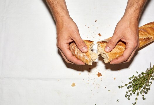 Top-down view of male hands breaking a fresh, crusty French baguette over a white linen tablecloth with scattered crumbs and a sprig of dried herbs.