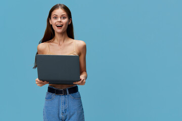 Obraz premium young woman holding laptop with surprised expression, casual clothing and blue background, technology and digital concept in modern lifestyle