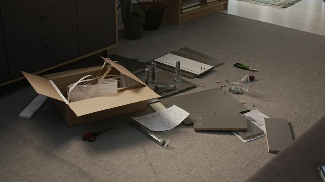 Full no people shot of parts of flatpack cabinet lying scattered around living room floor during assembly process, with opened box, fiberboard panels, fixtures, hardware, tools, instruction leaflet