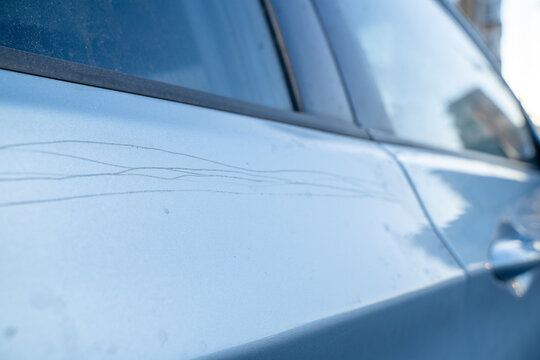 Closeup of a blue car door panel with long thin scratches across the paint, shallow depth of field showing vehicle bodywork damage, scuffs and reflections on metallic surface near the window trim edge