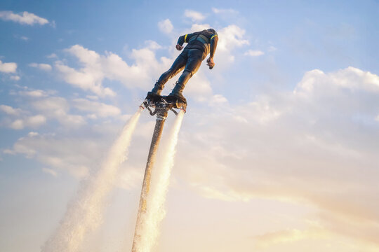 Person enjoys an exhilarating flyboarding experience above clear water, utilizing powerful jets to soar high into the sky. The bright sunlight casts a warm hue on the scene while scattered clouds