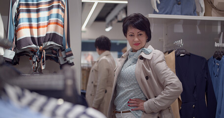 Woman trying on trench coat in fashion clothing store and looking to the camera