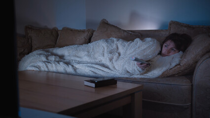 Woman Watching TV at Night Wrapped in Blanket on Couch, switches channels with the remote control