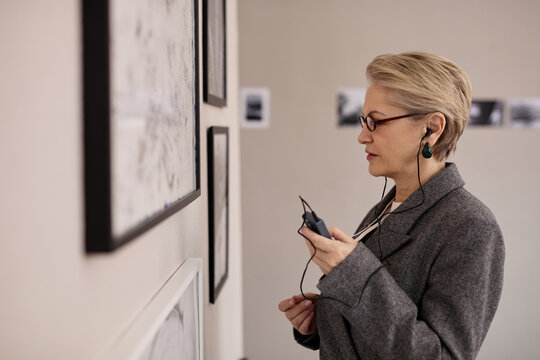 Middle aged Caucasian woman wearing glasses listening to audio guide while examining framed artwork on gallery wall, holding device in hand, standing in profile, focused on exhibit
