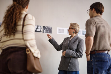 Middle aged Caucasian woman explaining black and white photographs on wall to young adult Caucasian man and woman during art discussion in gallery