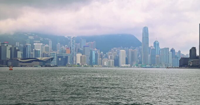 Panoramic view of Hong Kong Island skyscrapers from Victoria Harbour. Moody overcast sky over urban financial district. Iconic architecture and mountain backdrop. Hong Kong, 15 Apr 2025