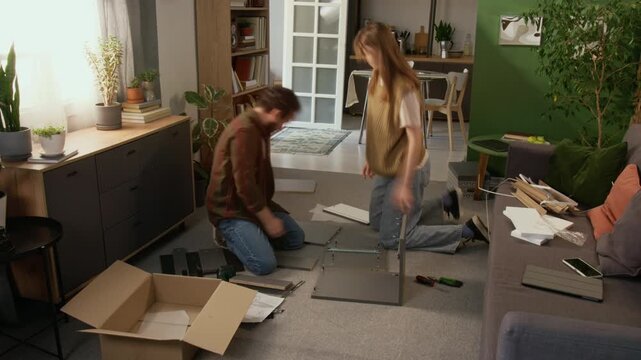 Wide high-angle timelapse shot of mature Caucasian couple unpacking box with furniture on floor, assembling cabinet from components and fixtures, working together and checking instructions