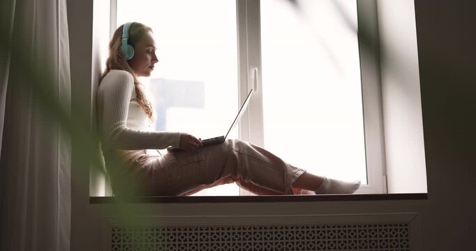 Young adult woman speaking on video call sits on window sill at home, using laptop for remote work, casual conversation with family. Counselling, online communication, videoconference application use
