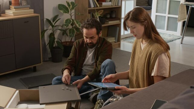 Full shot of middle-aged Caucasian woman with instructions on digital tablet assisting frustrated husband assembling flatpack cabinet at home, choosing fixtures and connectors from bag