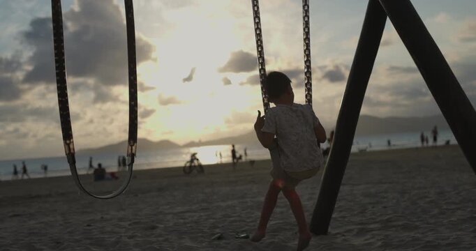Boy on swing set looking out towards sunset on beach - wide shot