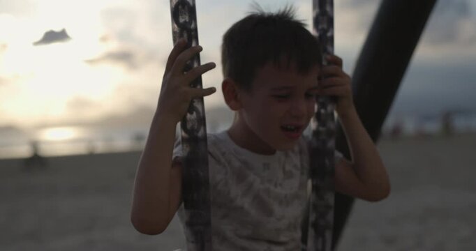 Toddler boy sits down on swing set on beach at sunset