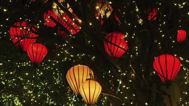 Hanging red and yellow lanterns illuminating a dark tree at night, creating an enchanting glow amidst strings of fairy lights for festive events and cultural celebrations