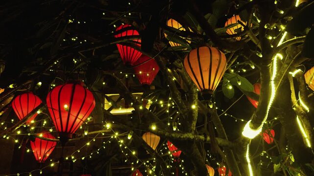 Hanging paper lanterns and string lights adorn a tree during the night, casting a warm and festive glow through the darkness, evoking a sense of tradition, celebration, and asian culture