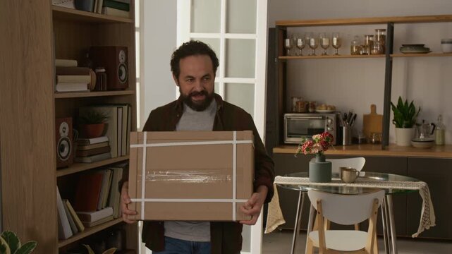 Medium tracking shot of joyous mature Caucasian man entering living room, carrying flatpack cardboard package with new furniture for assembly, then talking excitedly with wife and kissing her hand