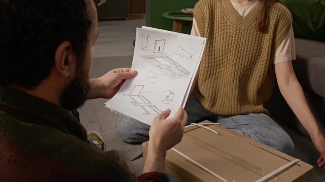 Medium over-shoulder shot of middle-aged bearded man studying assembly instructions while opening flat pack cardboard box with wife at home, to refurbish living room with new furniture
