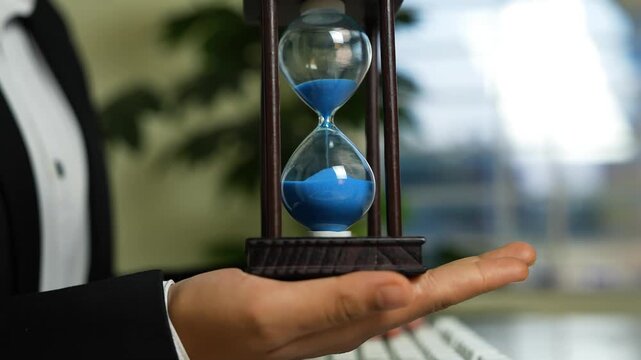 Hourglass with blue sand on hand, Hand holding a wooden-framed hourglass with blue sand, blurred office background