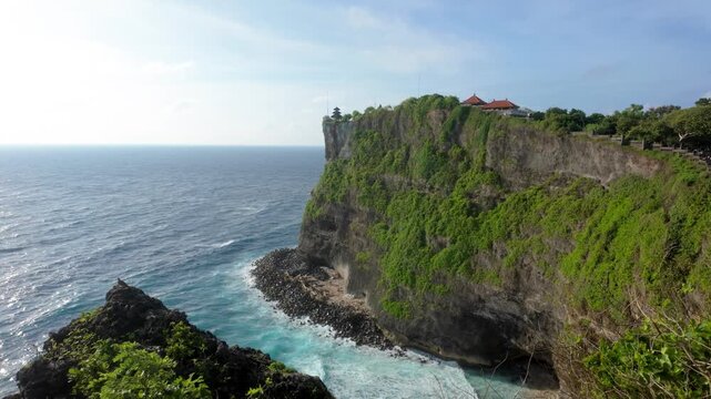 Uluwatu Temple, dramatic and epic cliff. Balinese Hindu temple located on the south-western tip of the Bukit Peninsula. Blue Ocean waves and sunshine. The camera elevates upwards to reveal the cliff. 