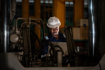 Industrial production worker wearing safety helmet using laptop computer between factory machines, smart manufacturing control and engineering technology in modern industry