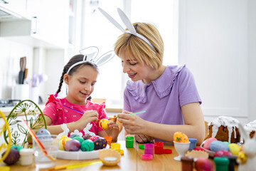 A mother and her daughter wearing bunny ears are happily painting colorful Easter eggs together at a kitchen table, surrounded by festive decorations and baked goods