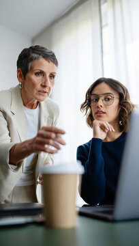 Veteran female executive intensely pinpointing a critical project flaw on the laptop screen while an attentive junior associate calmly processes the strict feedback.