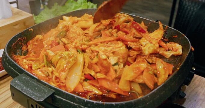 The process of cooking Korean-style spicy stir-fried spring chicken on an iron plate involves stir-frying tender chicken meat, chewy rice cakes, and kimchi in a bright red spicy sauce, releasing an en