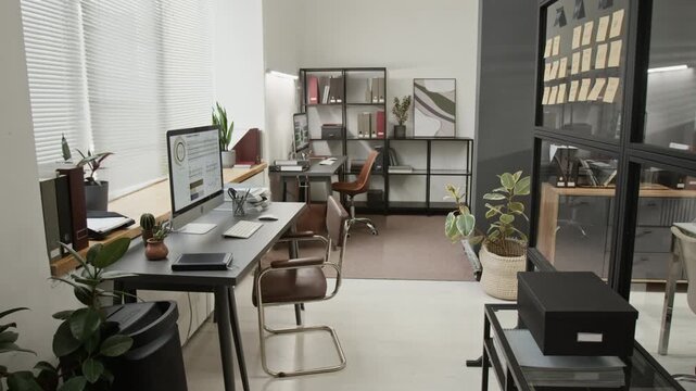 No people zoom in shot of modern well-organized office space with two work desks featuring desktop computers, comfortable chairs, potted plants and rack holding file folders