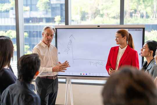 Senior businessman leading presentation with digital whiteboard in modern office. Meeting diverse creative team discussing marketing strategy success plan and corporate growth concept