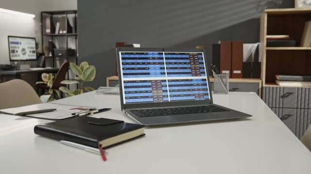 Close-up shot of white work table with laptop displaying spreadsheet, black-covered notebook, pens and applicants questionnaire attached to clipboard in modern office