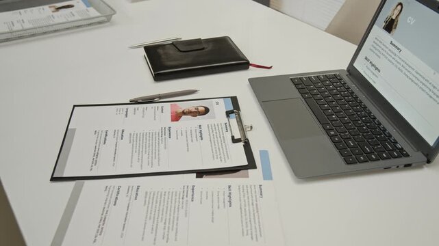 Close-up view of large office desk showing applicants resume on laptop screen, notebook, pens and printed questionnaires with photos