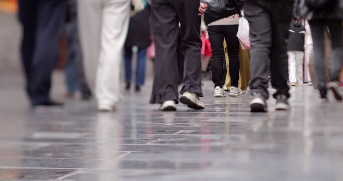 Low angle perspective of numerous people's legs and feet walking on a busy pedestrian street. Anonymous pedestrians moving along reflective stone pavement in an urban environment, slow motion shot.