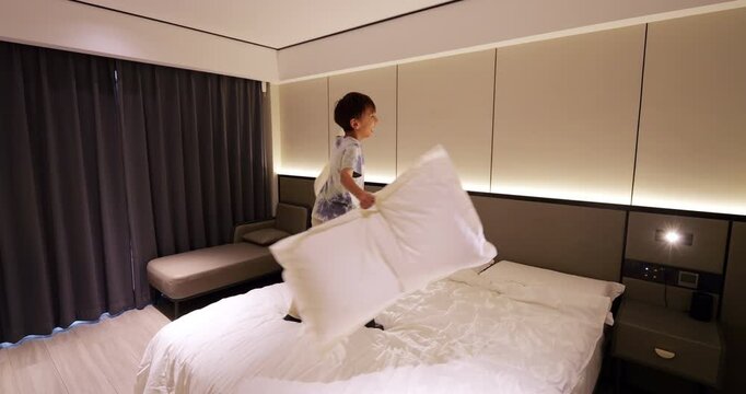 Slow motion shot of playful young boy having fun spinning and turning around with large pillows on king-size white bed in modern hotel room. He is smiling with mischievous and joyful expression