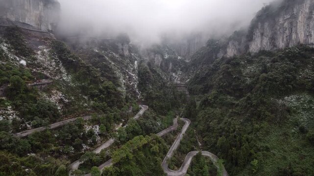 Aerial view of the curvy road leading up steep Tianmen Mountain in Zhangjiajie, steep rocky formations above in morning fog or clouds. Green plants and light dust of snow on the sheer slopes