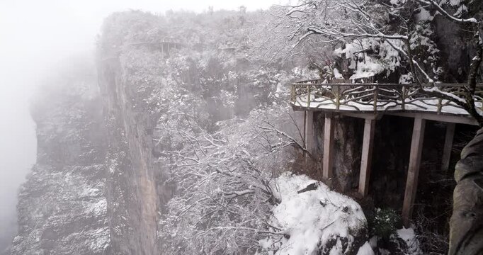Breathtaking high altitude trail along sheer mountain cliff in China's famous Tianmen Mountain park, with trees and pathways covered in fresh snow during cold, foggy day at end of March after storm