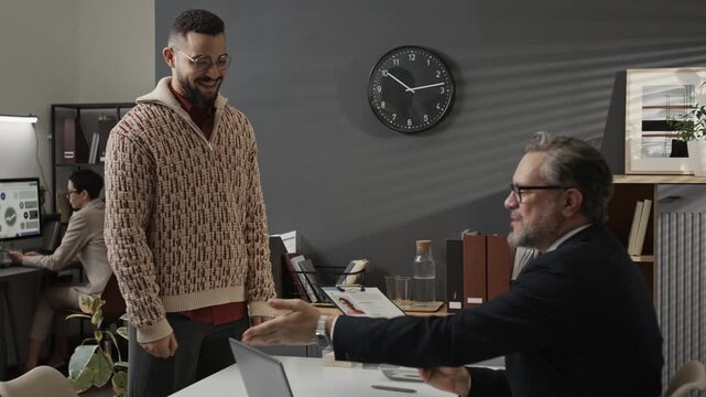 Zoom out view of gen Z applicant entering office and giving clipboard with questionnaire to middle-aged male recruiter inviting young man to take seat and starting conversation