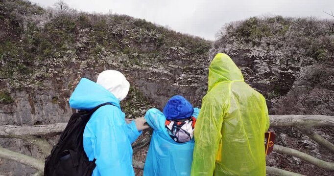 Happy tourist family with child wearing colorful raincoats stand at viewpoint over Heavens Gate, turn and lean down to look to famous cave from above. Camera glide forward and over to show view