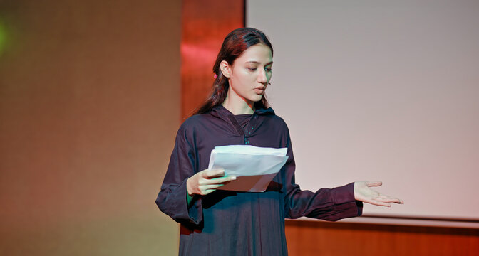 Indian young adult gen z female lady standing wear outfit dress hold script paper do play scene practice indoor hall stage. Teen age girl read story line talk speak day time college campus room place