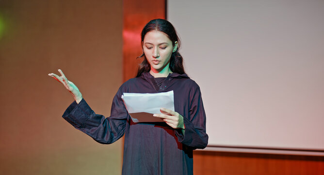 Indian young adult gen z female lady standing wear outfit dress hold script paper do play scene practice indoor hall stage. Teen age girl read story line talk speak day time college campus room place