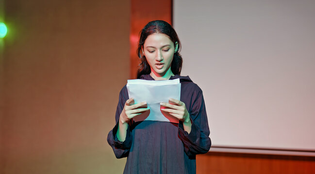 Indian young adult gen z female lady standing wear outfit dress hold script paper do play scene practice indoor hall stage. Teen age girl read story line talk speak day time college campus room place