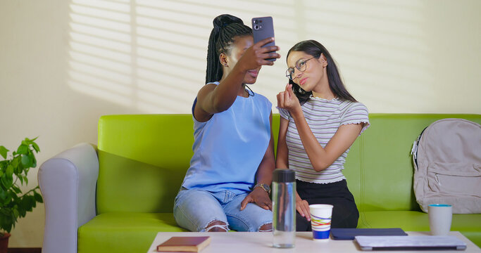 Happy young adult gen z woman sitting on sofa couch use mobile look cam take selfie indoor lounge room place teen age girl hold phone record casual video spend break time do funny pose enjoy new day