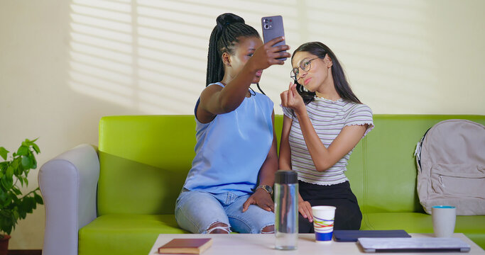 Happy young adult gen z woman sitting on sofa couch use mobile look cam take selfie indoor lounge room place teen age girl hold phone record casual video spend break time do funny pose enjoy new day