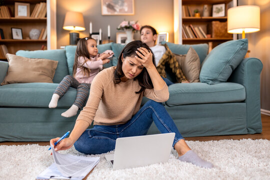 Stressed mother working on laptop while children play. Tired woman holding head with papers on floor. Work from home challenges and burnout concept. Busy parent trying to focus with noisy kids.