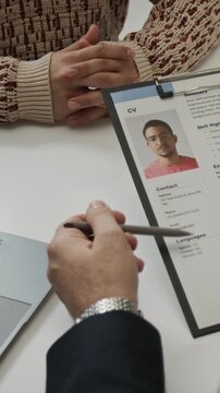 Vertical shot of unknown recruiter looking through resume of positive young man and speaking with him while conducting job interview in office