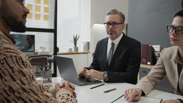 Elegant female hiring manager pointing at resume discussing it with gen Z applicant, her male colleague watching them and taking notes on laptop