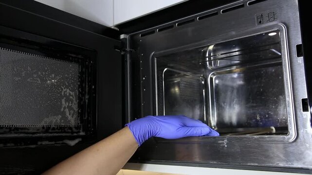 Person wearing blue latex gloves diligently cleans microwave oven interior. Grime and food residue removed from stainless steel surfaces for appliance hygiene.