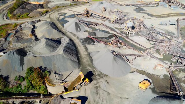 Aerial orbit of quarry with gravel stockpiles and processing conveyors, dump truck emptying fill into hopper
